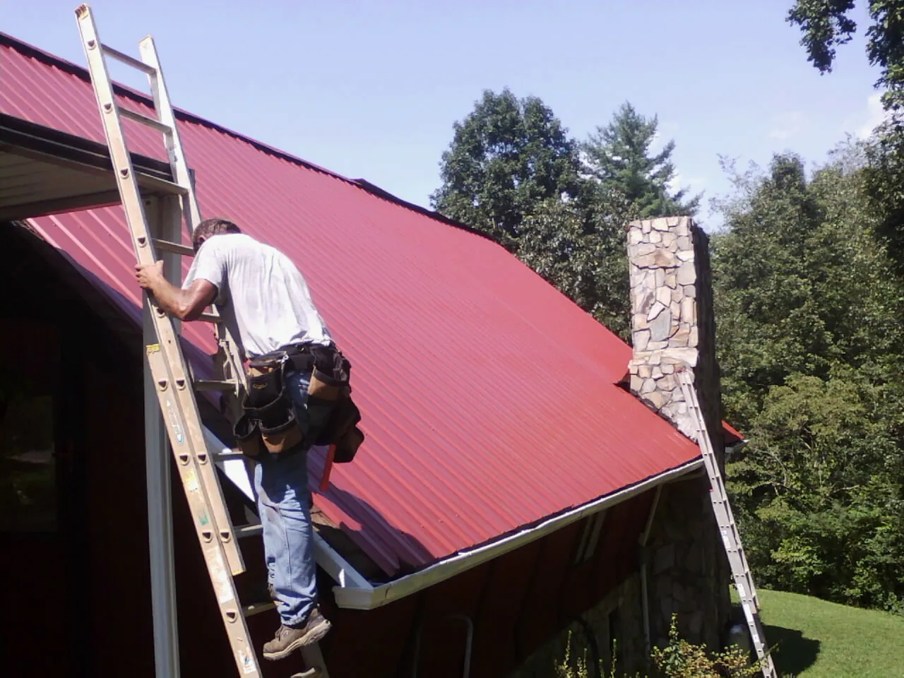 Roofer on a ladder beside a red metal roof and stone chimney — Soffit & Fascia Repair by Roofing Brothers in North Salem